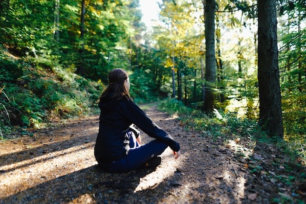A person sitting cross-legged in a forest, hands resting gently on their knees, meditating amidst the trees and dappled sunlight.