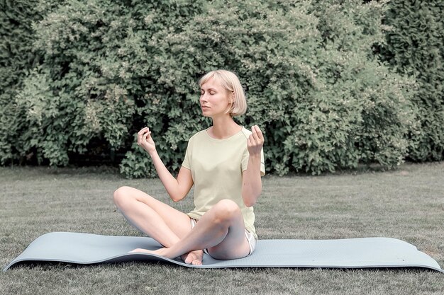 A peaceful scene in a park with a person demonstrating the seated position for body scan meditation. The person is sitting cross-legged on a yoga mat with hands resting gently on their knees.