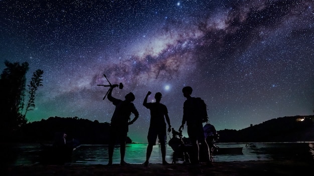 A panoramic night photograph showing a group of people looking up at the sky with telescopes and cameras set up. The Milky Way is brightly visible overhead, stretching across the sky.