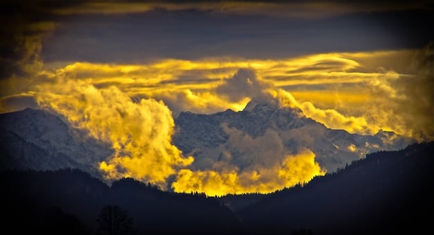 A wide shot of a mountain range at sunset, with the sun casting a golden glow on the peaks. Clouds are scattered across the sky, creating a dramatic and awe-inspiring scene.