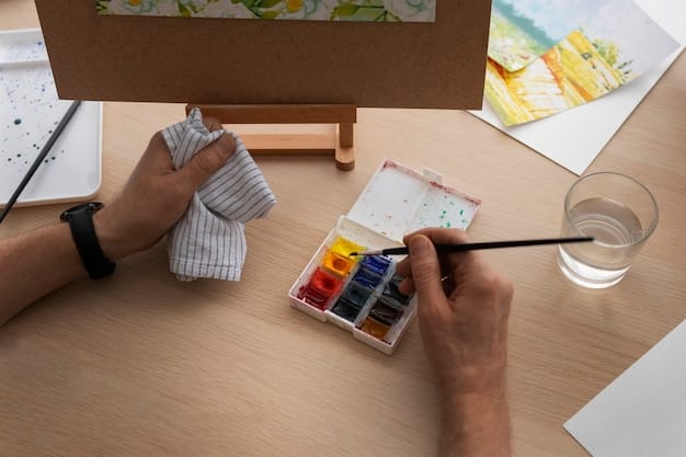 An overhead shot of a person painting with watercolor on a canvas. The person's hands are visible, holding a paintbrush and mixing colors on a palette. The background shows a variety of art supplies, such as brushes, paints, and jars of water.
