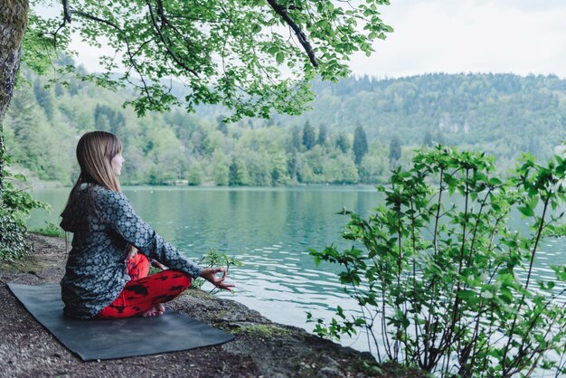 A woman practicing yoga outdoors with a view of mountains and a lake. She is holding a pose and repeating affirmations in her mind, looking peaceful and serene. The scene has a sense of tranquility and connection with nature.